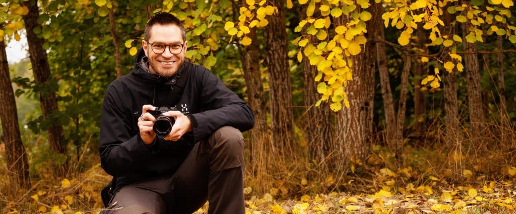 David Brohede med sin spegellösa systemkamera på huk framför höstträd.