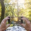 Bild på händer och en mobil som fotar mot solen i en skog