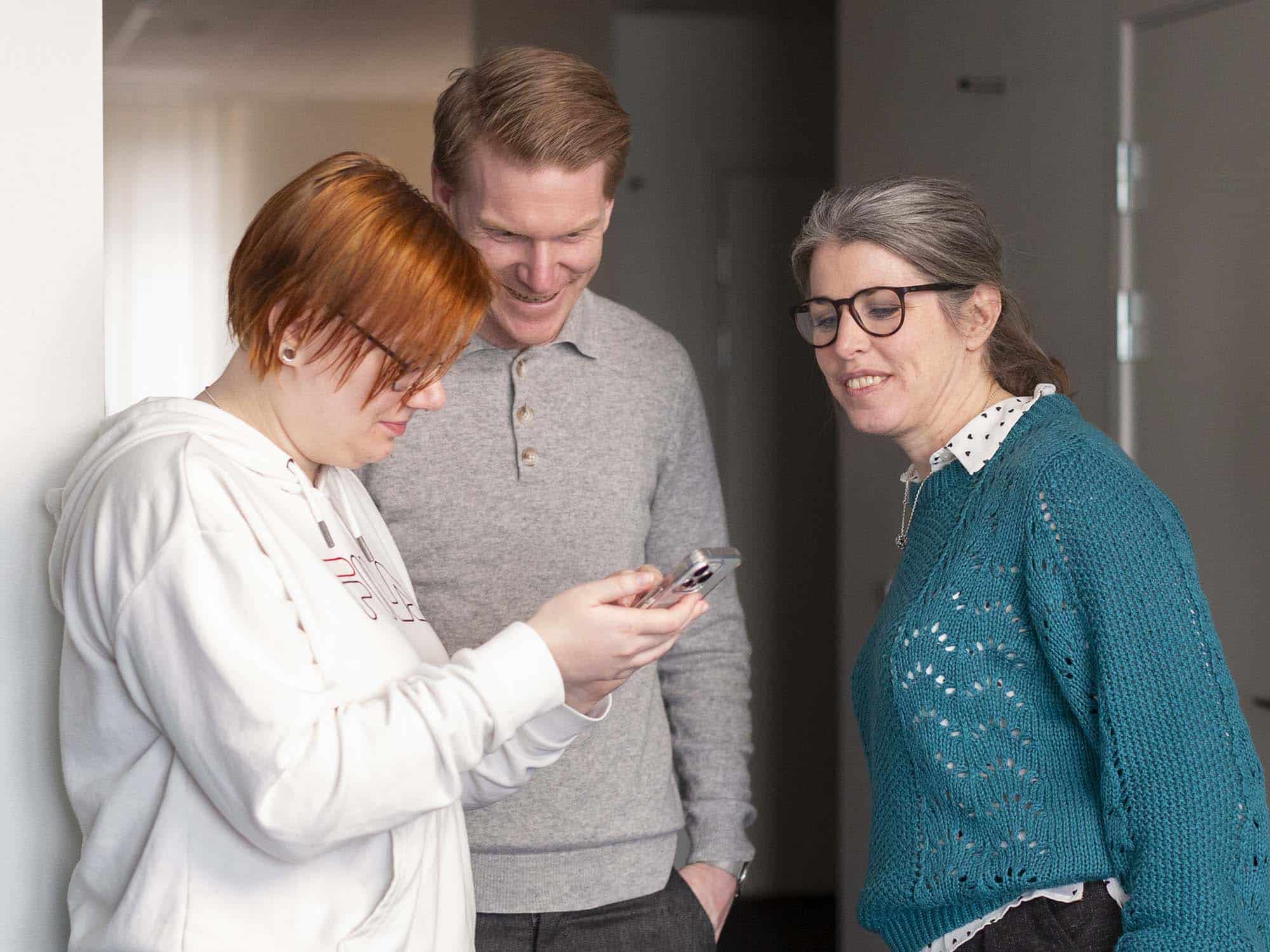 Tre personer ler och tittar tillsammans på skärmen på en mobiltelefon.