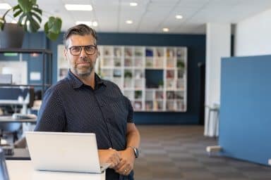 Patrik Gunnarsson, lärare i kursen Teams för projektledaren, står vid sin laptop och lutar sig mot en bänk i en kontorsmiljö.