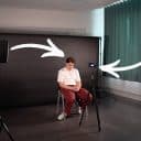 En person sitter på en stol framför en svart bakgrund i en fotostudio. En kamera är placerad på ett stativ framför personen. Två stora pilar pekar mot personen från två ljuskällor på stativ, en från vänster och en från höger. En fotograf justerar ljuset på vänster sida av bilden.