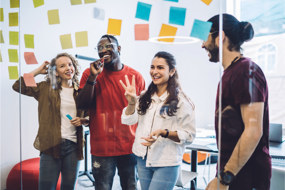 Fyra personer står tillsammans i ett modernt kontorsrum och samarbetar framför en glasvägg täckt med färgglada post-it-lappar. De skrattar och diskuterar idéer i en kreativ workshopmiljö. Ljuset är ljust och stämningen energisk och positiv.