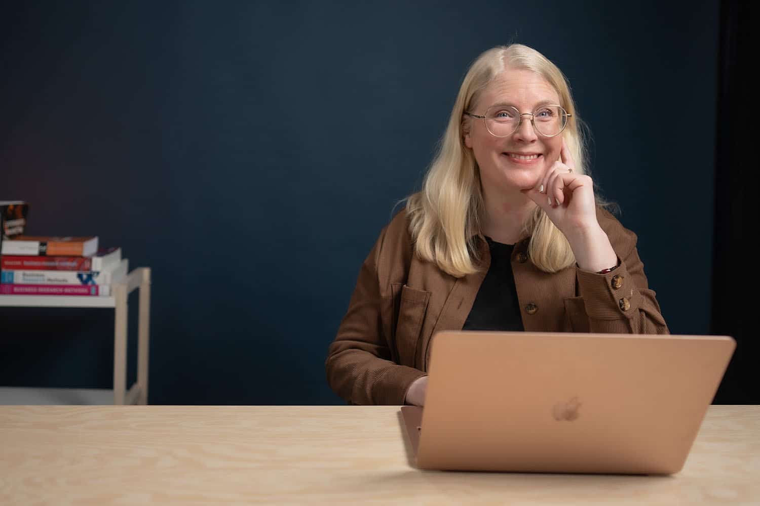 En glad Ida Serneberg Wallin med långt blont hår och glasögon sitter vid ett bord med en laptop framför sig. Hon lutar huvudet mot handen och tittar mot kameran. I bakgrunden syns en bokhylla med färgglada böcker mot en mörk blå vägg.
